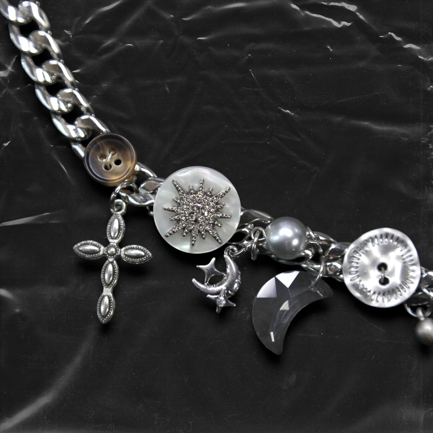 Chockychoker "Eclipsed Garden" choker features a silver chain base adorned with vintage-style brown and metallic buttons, an engraved rose pendant, faux pearls, smoky resin beads, moon-and-star charms, a silver cross, a button decorated with a white, snowflake-shaped crystal inlay, and a long silver spike pendant. Blending dreamy and retro aesthetics, this design evokes a romantic fairytale meets Y2K mood perfect for those who seek storytelling accessories and distinctive personal style.