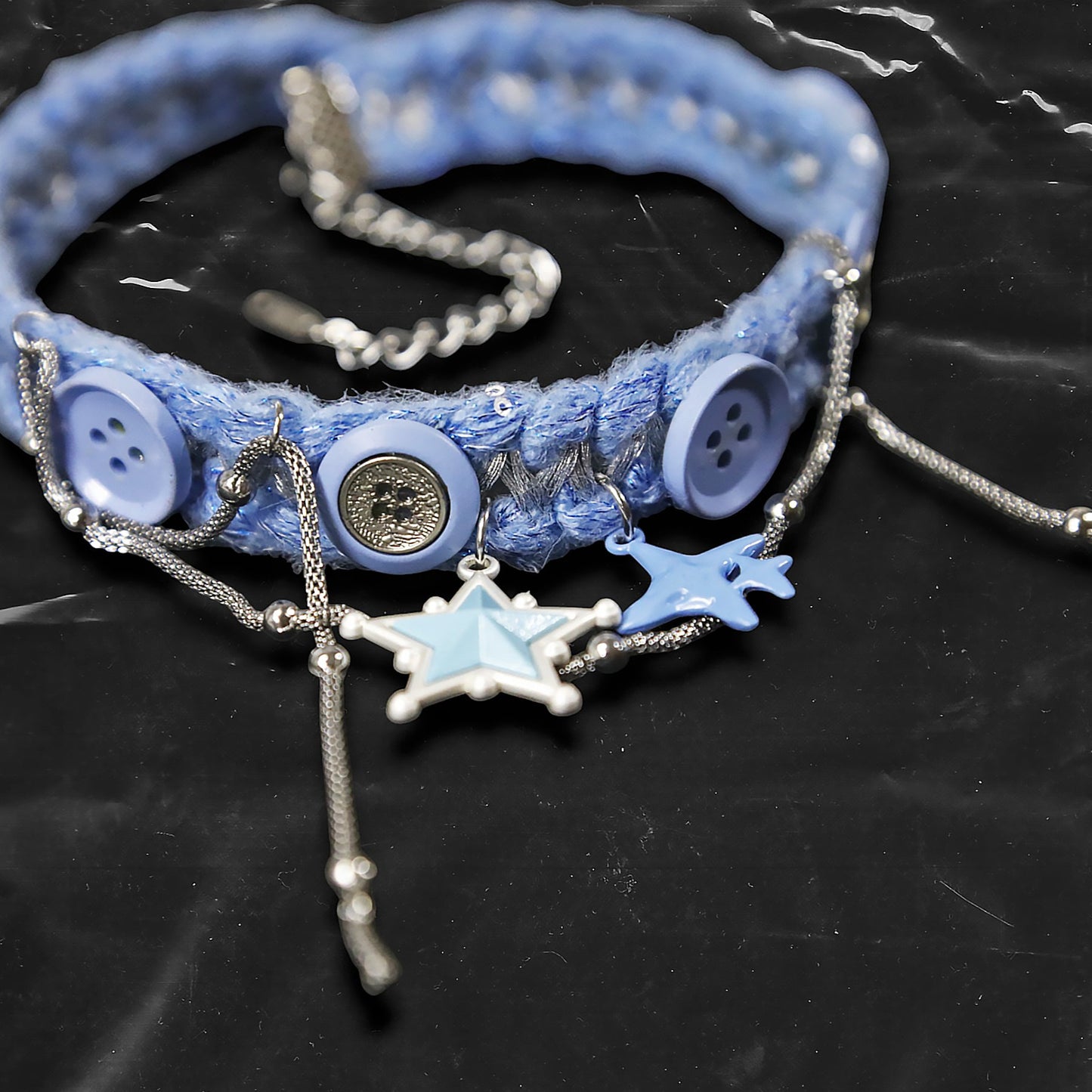 Chockychoker "Blue Save Point" handmade choker featuring blue star pendants in resin finish, accented with silver-tone chains of mixed texture, and secured onto a woven light blue yarn base. Decorated with multiple blue and silver-tone buttons along the band. This piece combines star motifs , creating a unique blend of sweet-cool , alt, Harajuku, cyberpop, and soft punk aesthetics. Materials include yarn, alloy buttons, silver-tone metal chains, and enamel resin charms.