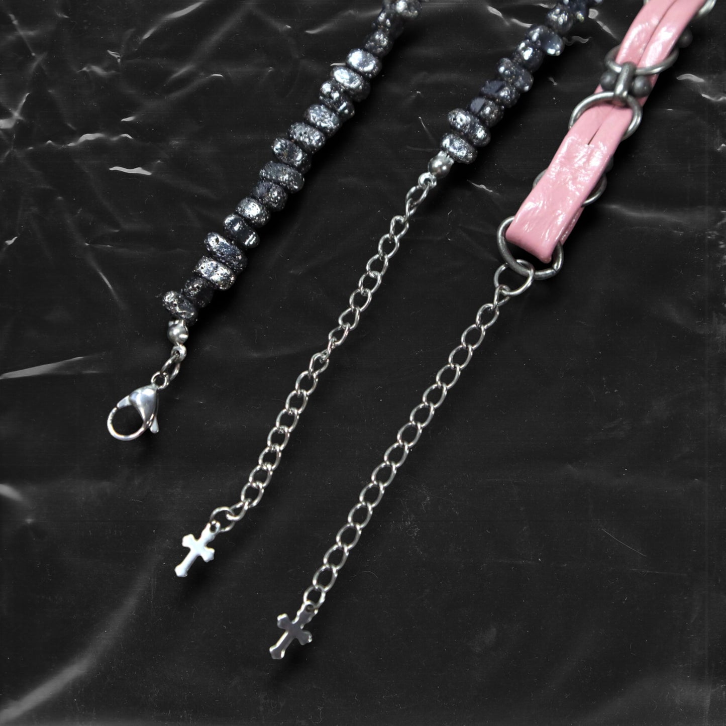 Chockychoker “Angel Trap” is a handcrafted dual-layer choker combining a braided pink faux leather strap interwoven with black-silver alloy components on the top layer, accented by a silver metal cross pendant. The lower layer features chunky metal beads with embossed, textured surfaces and a silver-tone cross charm embedded with light pink crystal-like stones. Multiple dangling silver chains add dimension and movement. This statement accessory making it ideal for Y2K, cyber-goth, punk,  and  rich materials