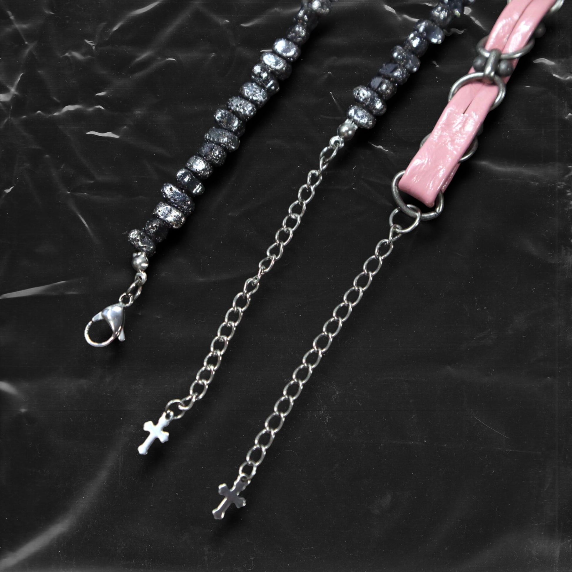 Chockychoker “Angel Trap” is a handcrafted dual-layer choker combining a braided pink faux leather strap interwoven with black-silver alloy components on the top layer, accented by a silver metal cross pendant. The lower layer features chunky metal beads with embossed, textured surfaces and a silver-tone cross charm embedded with light pink crystal-like stones. Multiple dangling silver chains add dimension and movement. This statement accessory making it ideal for Y2K, cyber-goth, punk,  and  rich materials