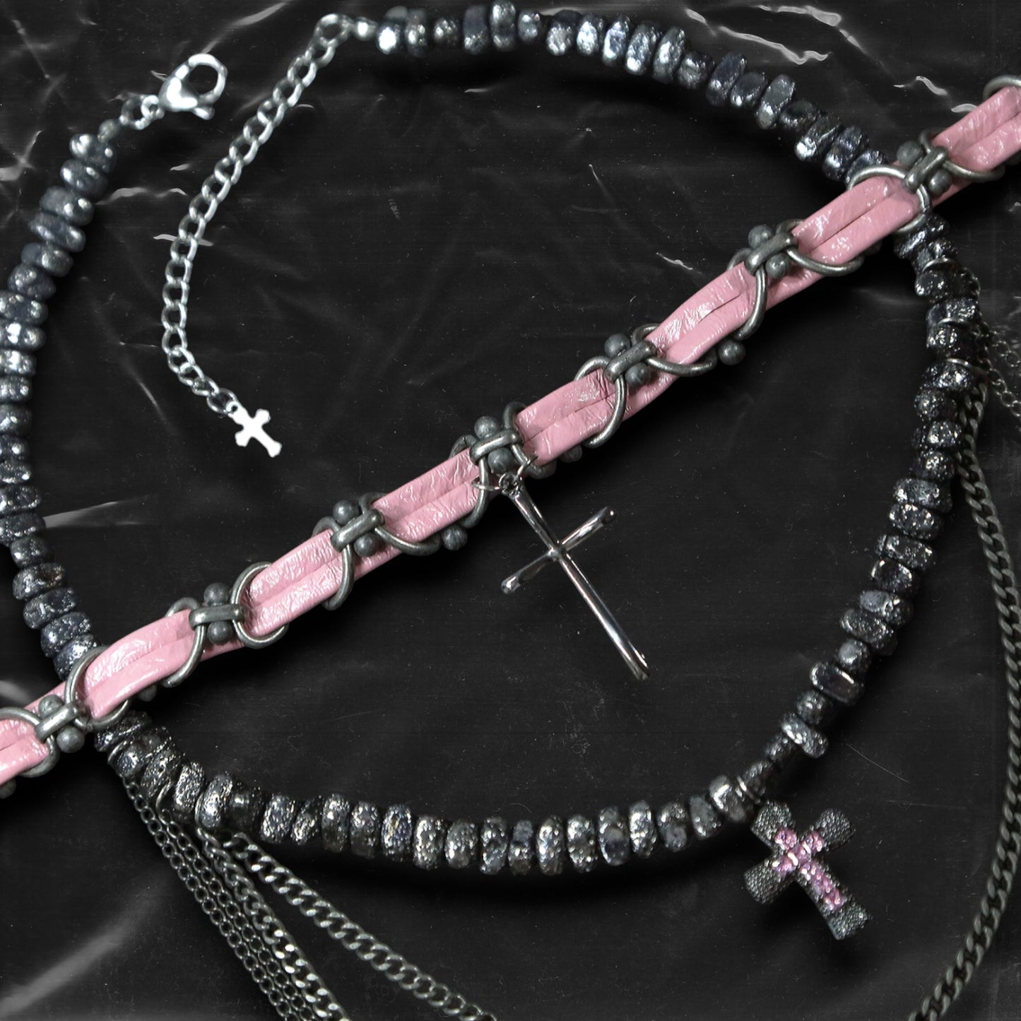 Chockychoker “Angel Trap” is a handcrafted dual-layer choker combining a braided pink faux leather strap interwoven with black-silver alloy components on the top layer, accented by a silver metal cross pendant. The lower layer features chunky metal beads with embossed, textured surfaces and a silver-tone cross charm embedded with light pink crystal-like stones. Multiple dangling silver chains add dimension and movement. This statement accessory making it ideal for Y2K, cyber-goth, punk,  and  rich materials