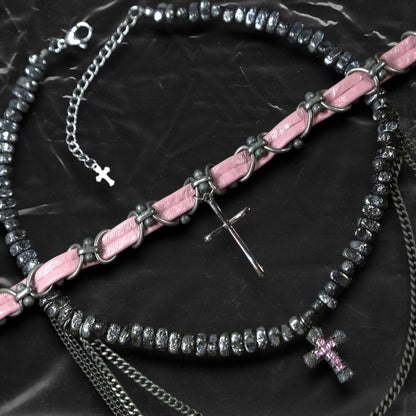 Chockychoker “Angel Trap” is a handcrafted dual-layer choker combining a braided pink faux leather strap interwoven with black-silver alloy components on the top layer, accented by a silver metal cross pendant. The lower layer features chunky metal beads with embossed, textured surfaces and a silver-tone cross charm embedded with light pink crystal-like stones. Multiple dangling silver chains add dimension and movement. This statement accessory making it ideal for Y2K, cyber-goth, punk,  and  rich materials