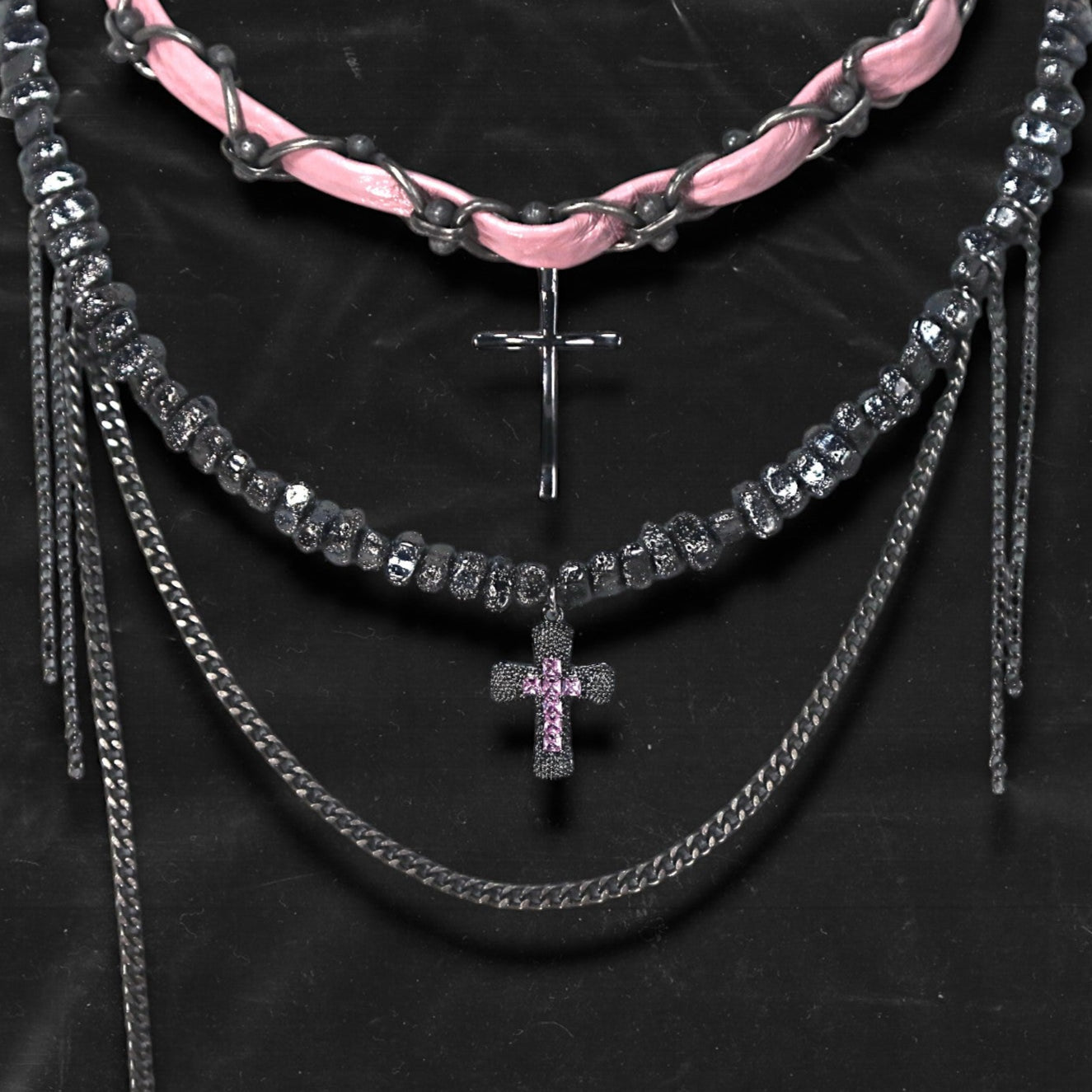 Chockychoker “Angel Trap” is a handcrafted dual-layer choker combining a braided pink faux leather strap interwoven with black-silver alloy components on the top layer, accented by a silver metal cross pendant. The lower layer features chunky metal beads with embossed, textured surfaces and a silver-tone cross charm embedded with light pink crystal-like stones. Multiple dangling silver chains add dimension and movement. This statement accessory making it ideal for Y2K, cyber-goth, punk,  and  rich materials