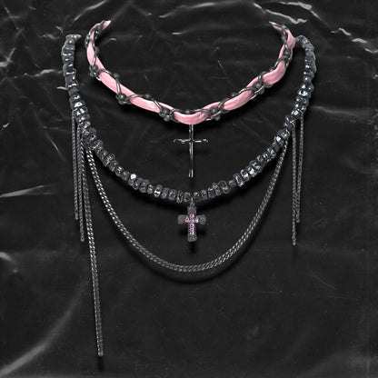 Chockychoker “Angel Trap” is a handcrafted dual-layer choker combining a braided pink faux leather strap interwoven with black-silver alloy components on the top layer, accented by a silver metal cross pendant. The lower layer features chunky metal beads with embossed, textured surfaces and a silver-tone cross charm embedded with light pink crystal-like stones. Multiple dangling silver chains add dimension and movement. This statement accessory making it ideal for Y2K, cyber-goth, punk,  and  rich materials