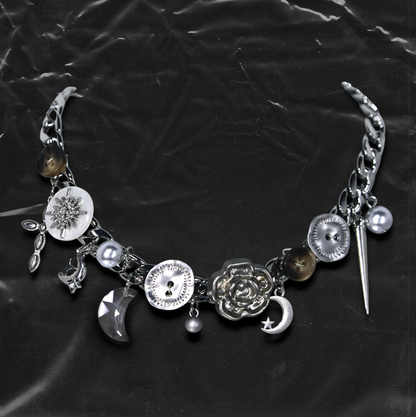 Chockychoker "Eclipsed Garden" choker features a silver chain base adorned with vintage-style brown and metallic buttons, an engraved rose pendant, faux pearls, smoky resin beads, moon-and-star charms, a silver cross, a button decorated with a white, snowflake-shaped crystal inlay, and a long silver spike pendant. Blending dreamy and retro aesthetics, this design evokes a romantic fairytale meets Y2K mood perfect for those who seek storytelling accessories and distinctive personal style.