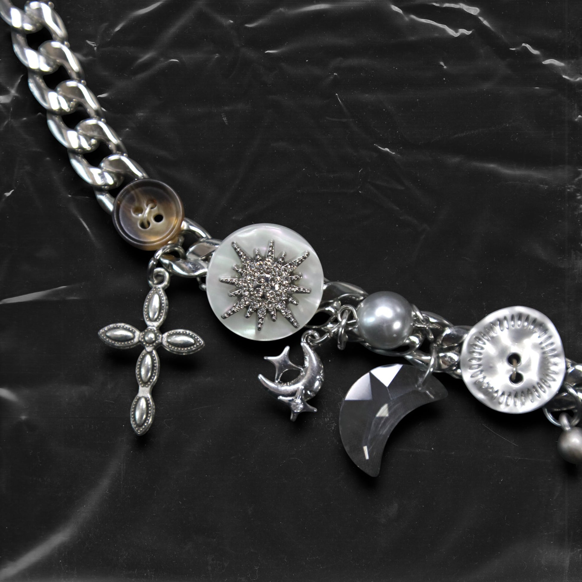 Chockychoker "Eclipsed Garden" choker features a silver chain base adorned with vintage-style brown and metallic buttons, an engraved rose pendant, faux pearls, smoky resin beads, moon-and-star charms, a silver cross, a button decorated with a white, snowflake-shaped crystal inlay, and a long silver spike pendant. Blending dreamy and retro aesthetics, this design evokes a romantic fairytale meets Y2K mood perfect for those who seek storytelling accessories and distinctive personal style.