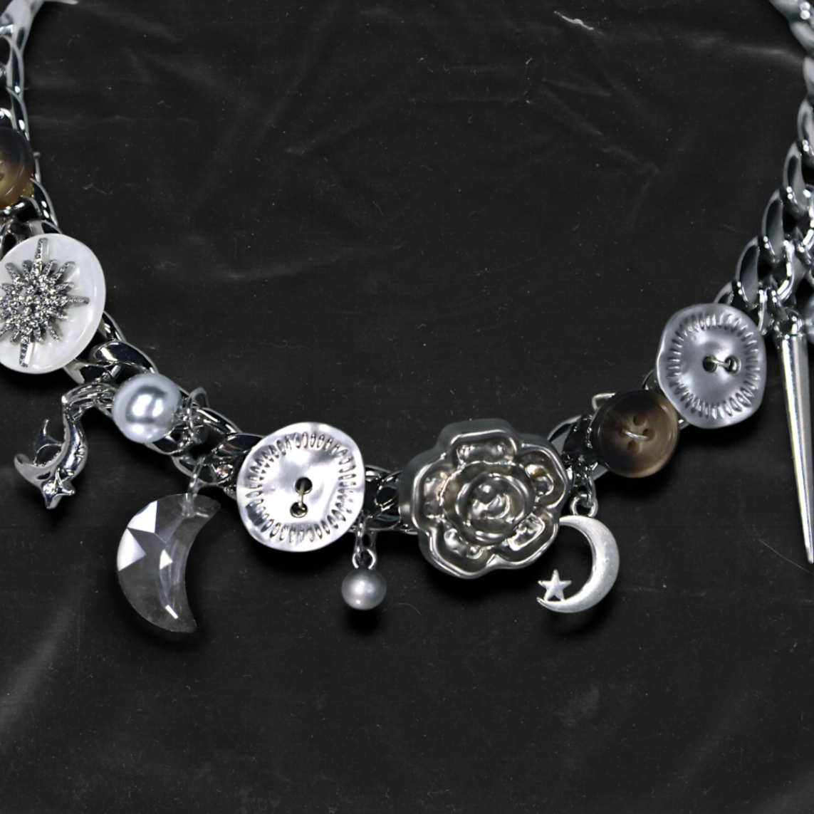 Chockychoker "Eclipsed Garden" choker features a silver chain base adorned with vintage-style brown and metallic buttons, an engraved rose pendant, faux pearls, smoky resin beads, moon-and-star charms, a silver cross, a button decorated with a white, snowflake-shaped crystal inlay, and a long silver spike pendant. Blending dreamy and retro aesthetics, this design evokes a romantic fairytale meets Y2K mood perfect for those who seek storytelling accessories and distinctive personal style.