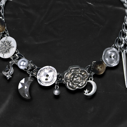 Chockychoker "Eclipsed Garden" choker features a silver chain base adorned with vintage-style brown and metallic buttons, an engraved rose pendant, faux pearls, smoky resin beads, moon-and-star charms, a silver cross, a button decorated with a white, snowflake-shaped crystal inlay, and a long silver spike pendant. Blending dreamy and retro aesthetics, this design evokes a romantic fairytale meets Y2K mood perfect for those who seek storytelling accessories and distinctive personal style.