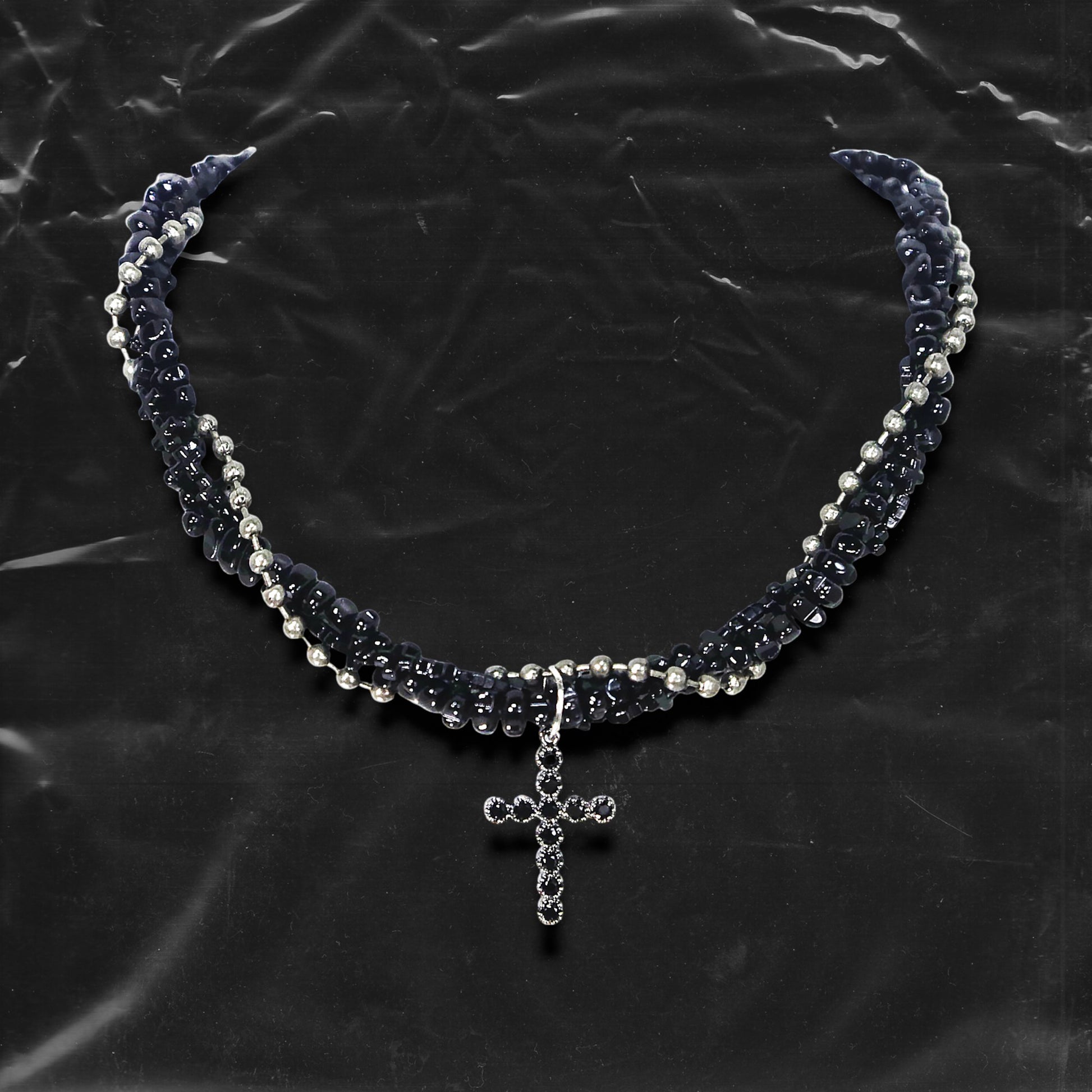Chockychoker “Midnight Stitch” choker features a layered design made of black beads and silver-tone faux pearls, centered with a black rhinestone-studded cross pendant. The necklace blends gothic and Y2K influences with bold contrast and structured detail. A perfect statement piece for gothic, punk, alt, emo, and fashion-forward styles.