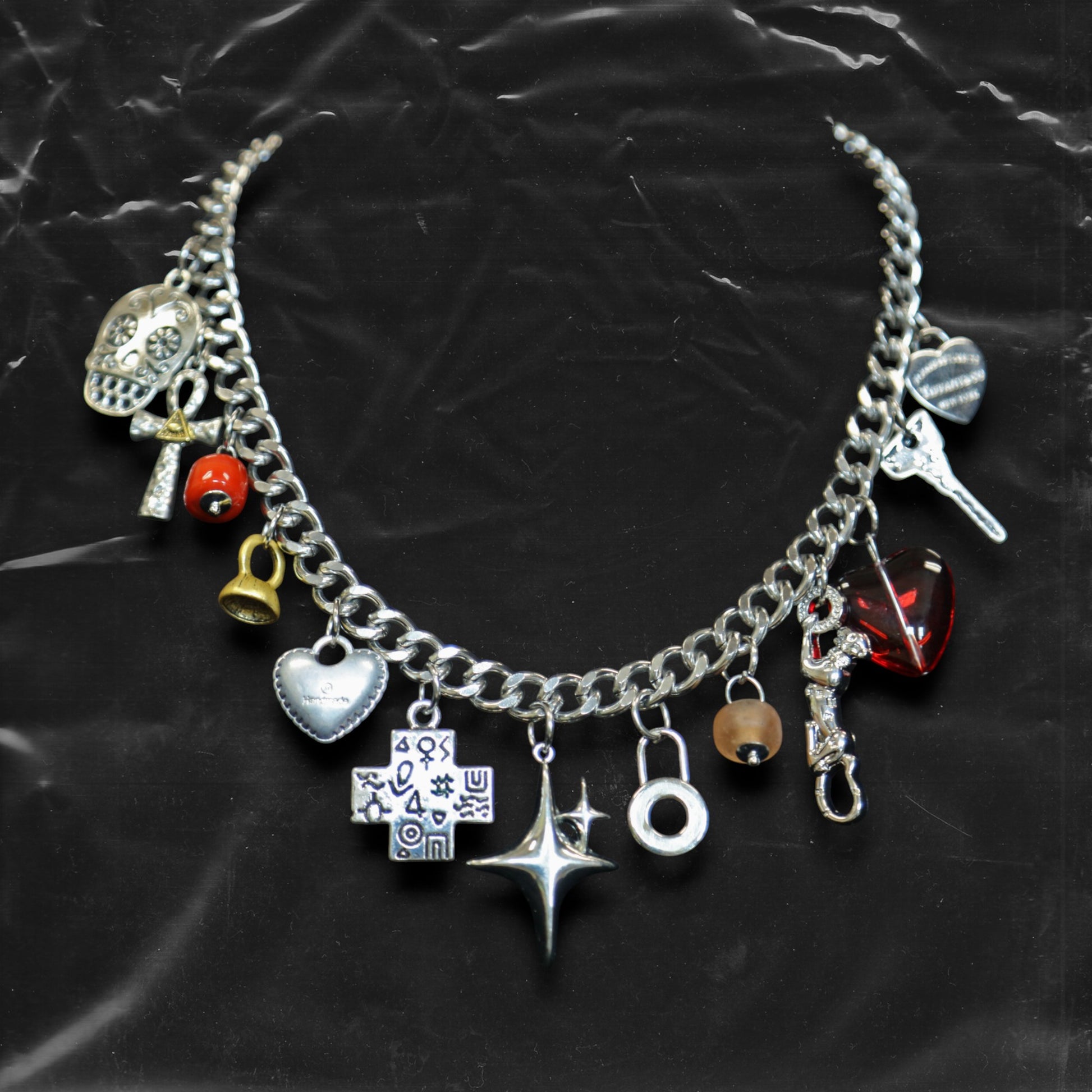 Chockychoker "Moonlit Relic" choker featuring a thick silver-tone chain adorned with eclectic dangling charms, including a crystal-eyed skull, metallic hearts, engraved cross, cosmic star, abstract rings, red gem heart, and vintage-style miniatures like a bell, shell, and keys. This mixed-media statement piece blends Y2K, gothic, alt, and symbolic aesthetics for bold and expressive styling.
