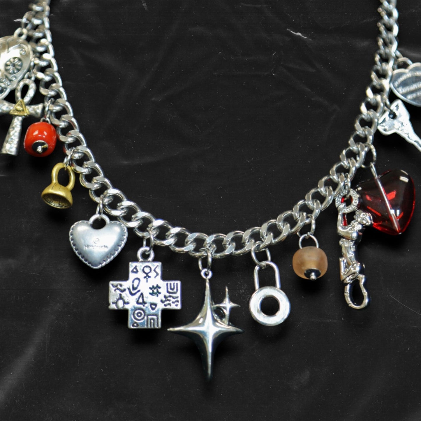 Chockychoker "Moonlit Relic" choker featuring a thick silver-tone chain adorned with eclectic dangling charms, including a crystal-eyed skull, metallic hearts, engraved cross, cosmic star, abstract rings, red gem heart, and vintage-style miniatures like a bell, shell, and keys. This mixed-media statement piece blends Y2K, gothic, alt, and symbolic aesthetics for bold and expressive styling.

