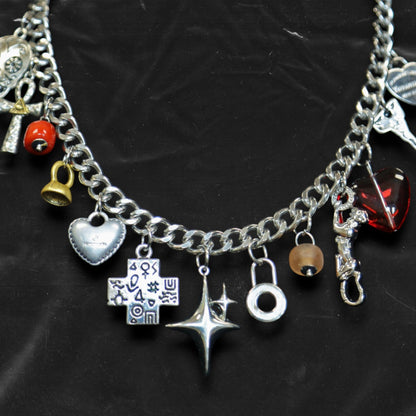 Chockychoker "Moonlit Relic" choker featuring a thick silver-tone chain adorned with eclectic dangling charms, including a crystal-eyed skull, metallic hearts, engraved cross, cosmic star, abstract rings, red gem heart, and vintage-style miniatures like a bell, shell, and keys. This mixed-media statement piece blends Y2K, gothic, alt, and symbolic aesthetics for bold and expressive styling.
