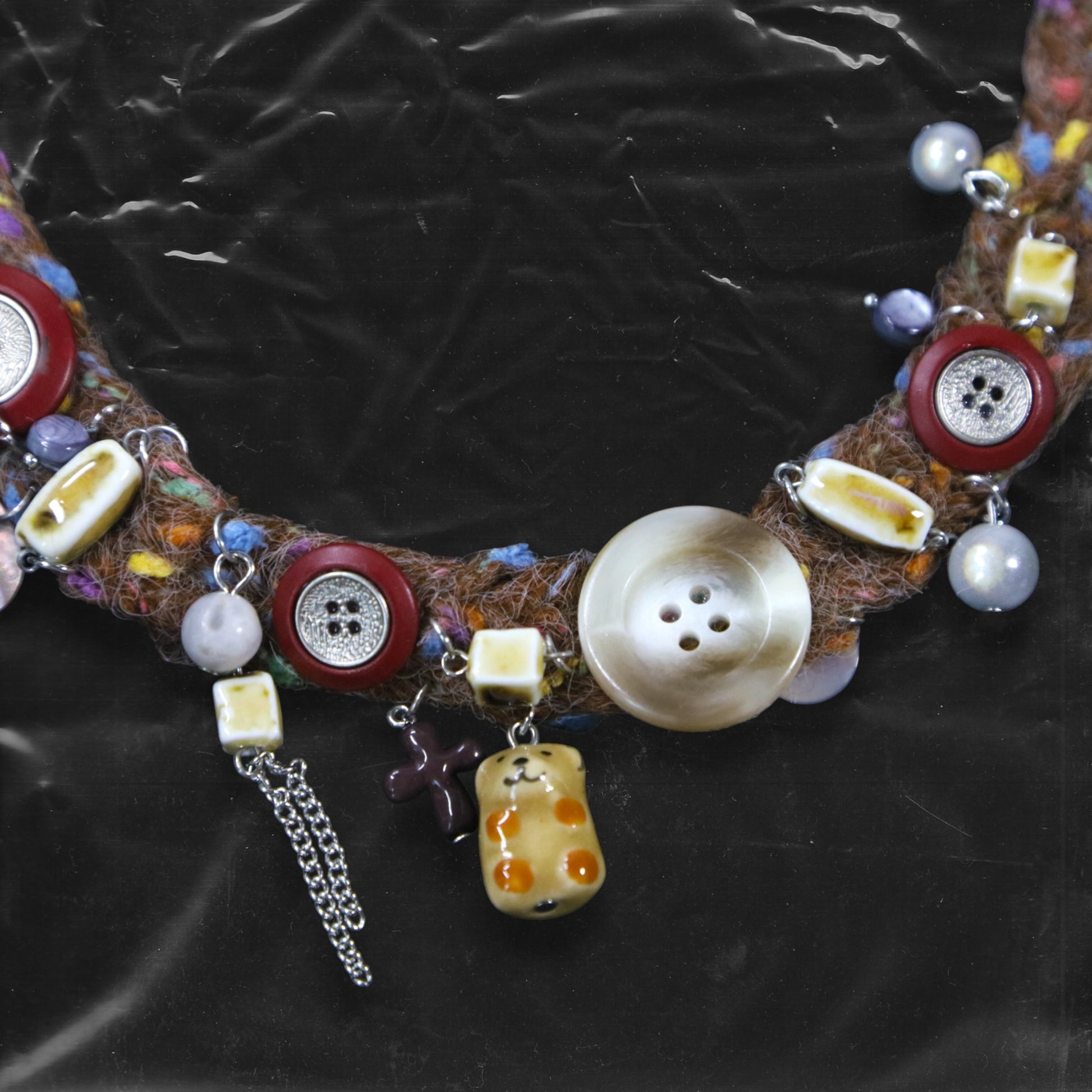 Chockychoker “Teddy Patch I” is a handcrafted choker made from colorful handwoven  yarn, featuring a central oversized iridescent brown button, ceramic bear charm, deep red ceramic cross pendant, shell accents, pearl-like beads, translucent resin blocks, layered silver-tone chains, metallic star charm, glossy red and mixed buttons, and scattered gemstone style plastic beads. Rich in textures and asymmetrical charm arrangement eclectic necklace embodies . Ideal for Y2K, gothic, Harajuku, grunge, retro,  boho