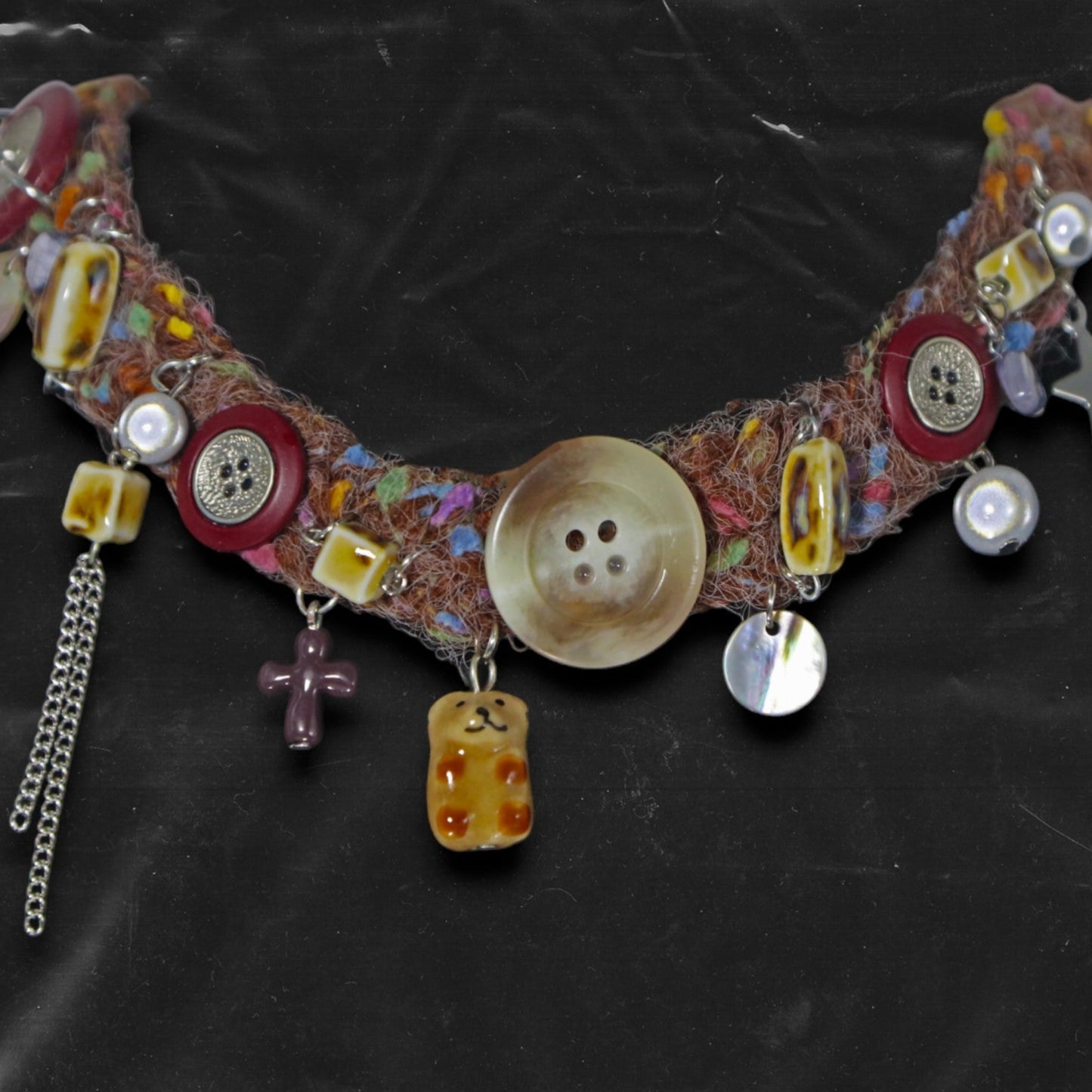 Chockychoker “Teddy Patch I” is a handcrafted choker made from colorful handwoven  yarn, featuring a central oversized iridescent brown button, ceramic bear chapendant, shell accents, pearl-like beads, translucent resin blocks, layered silver-tone chains, metallic star charm, glossy red and mixed buttons, and scattered gemstone-style plastic beads. Rich in textures and asymmetrical charm arrangement eclectic necklace embodies . Ideal for Y2K, gothic, indie, Harajuku, grunge, artsy, retro, alt,  boho fashion
