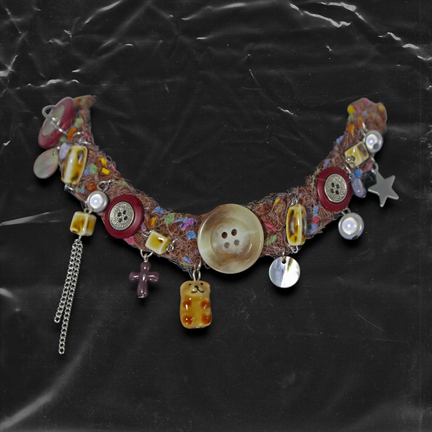 Chockychoker “Teddy Patch I” is a handcrafted choker made from colorful handwoven  yarn, featuring a central oversized iridescent brown button, ceramic bear charm, deep red ceramic cross pendant, shell accents, pearl-like beads, translucent resin blocks, layered silver-tone chains, metallic star charm, glossy red and mixed buttons, and scattered gemstone style plastic beads. Rich in textures and asymmetrical charm arrangement eclectic necklace embodies . Ideal for Y2K, gothic, Harajuku, grunge, retro,  boho