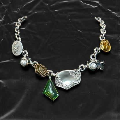 Chockychoker "Witch's Pocket" choker features a whimsical mix of charms, including sculpted silver-toned metal, a delicate milky-white button set with a snowflake-shaped faux crystal, a gold shell charm, faux pearls, a translucent green gemstone pendant, engraved medallions, and abstract vintage elements. The overall style blends eccentricity, fairycore, vintage, witchy, and Y2K aesthetics—perfect for those who embrace unique and expressive fashion.