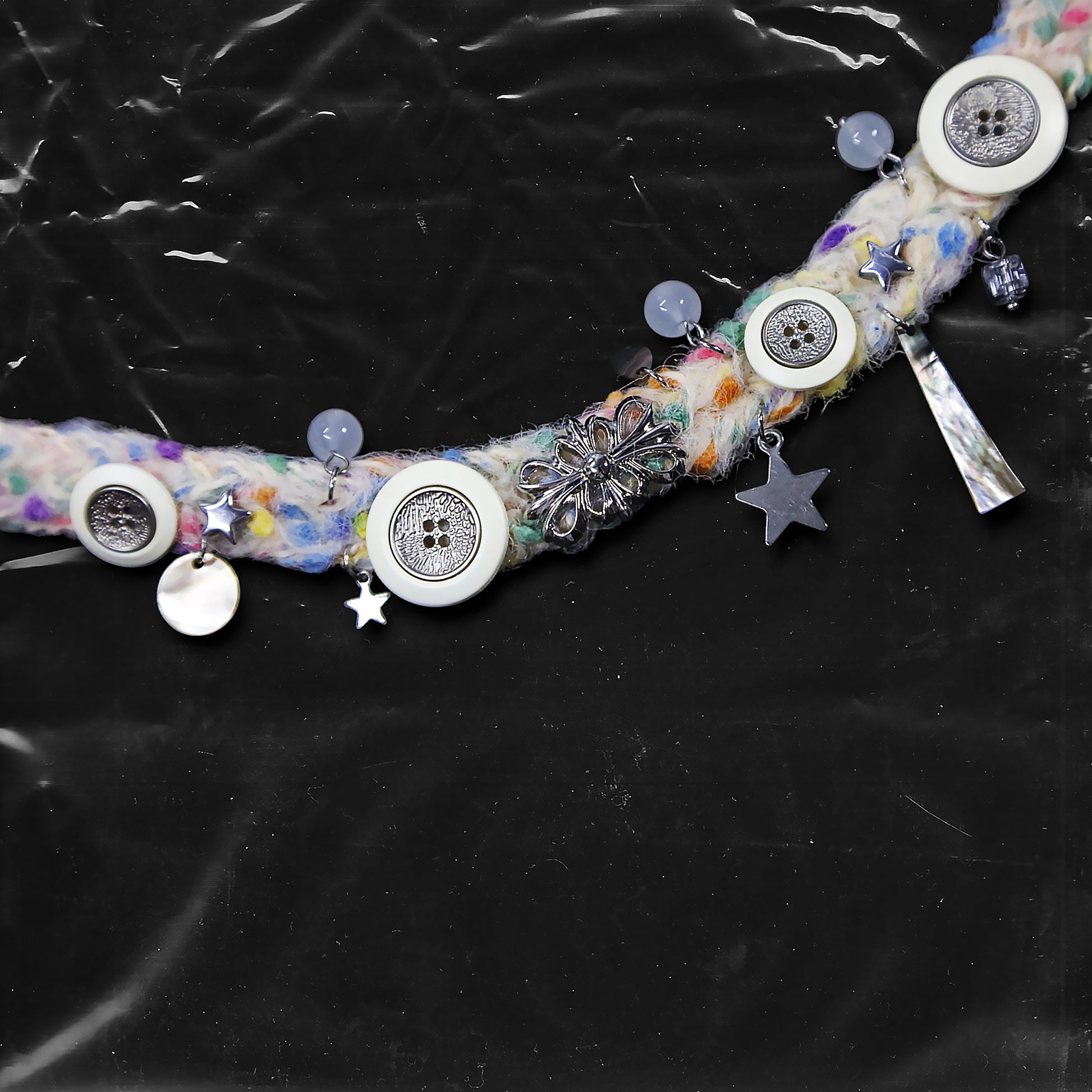 Chockychoker “Woolen Wish” is a handmade choker crafted from multicolored yarn with green, purple, yellow, and pink. Adorned with white and metallic vintage-style buttons, iridescent translucent beads, opalescent shell discs, and an ornate silver-tone floral centerpiece. Accents include soft textured fibers, dangling orb charms, and layered mixed materials that add depth and movement. A harmonious mix of fairycore, cottagecore, Y2K, and alternative aesthetic perfect for indie, vintage inspired, boho, artist