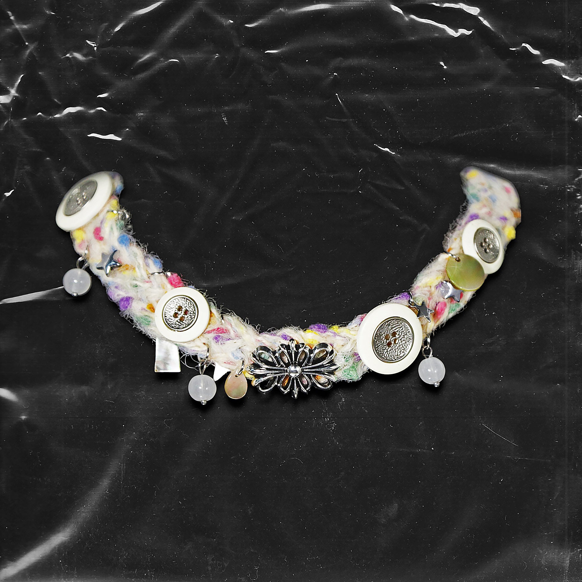 Chockychoker “Woolen Wish” is a handmade choker crafted from multicolored yarn with green, purple, yellow, and pink. Adorned with white and metallic vintage-style buttons, iridescent translucent beads, opalescent shell discs, and an ornate silver-tone floral centerpiece. Accents include soft textured fibers, dangling orb charms, and layered mixed materials that add depth and movement. A harmonious mix of fairycore, cottagecore, Y2K, and alternative aesthetic perfect for indie, vintage inspired, boho, artis
