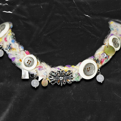 Chockychoker “Woolen Wish” is a handmade choker crafted from multicolored yarn with green, purple, yellow, and pink. Adorned with white and metallic vintage-style buttons, iridescent translucent beads, opalescent shell discs, and an ornate silver-tone floral centerpiece. Accents include soft textured fibers, dangling orb charms, and layered mixed materials that add depth and movement. A harmonious mix of fairycore, cottagecore, Y2K, and alternative aesthetic perfect for indie, vintage inspired, boho, artis
