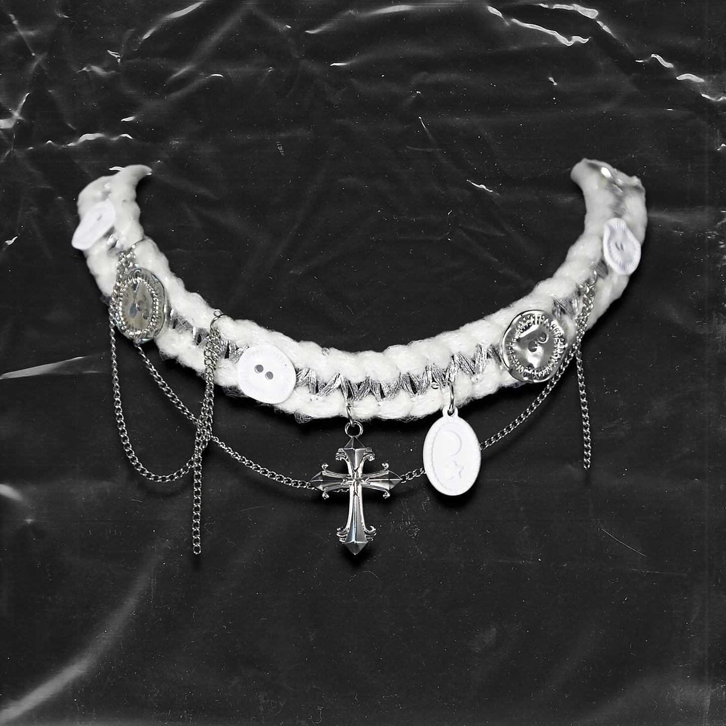 Chockychoker “Milk Galaxy” is a Handcrafted gothic-style choker made from soft white yarn interwoven with shimmering silver thread, adorned with layered silver-tone chains, a central baroque-style silver cross pendant, a mix of glossy white plastic buttons and vintage-style metallic buttons, and white oval resin charms ideal for lovers of gothic, alt, punk, lolita, soft grunge, and romantic street fashion. Perfect for those seeking statement chokers with unique handcrafted details and mixed media textures.