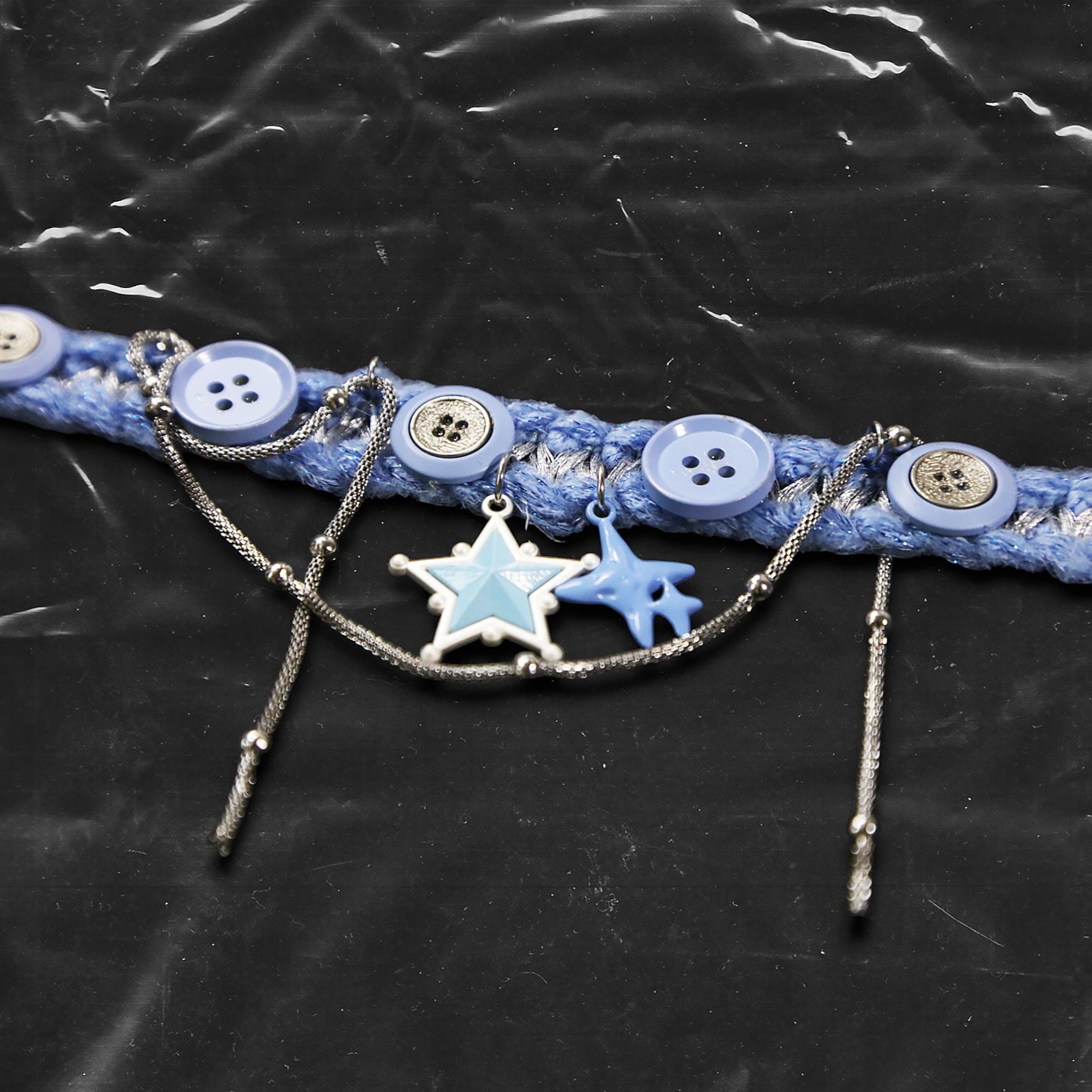Chockychoker "Blue Save Point"  handmade choker featuring  blue star pendants in resin   finish, accented with silver-tone chains of mixed texture, and secured onto a woven light blue yarn base. Decorated with multiple blue and silver-tone buttons along the band. This piece combines  star motifs , creating a unique blend of sweet-cool , alt, Harajuku, cyberpop, and soft punk aesthetics.  Materials include yarn, alloy buttons, silver-tone metal chains, and enamel resin charms.
